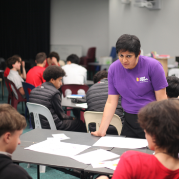 Woman in a Local Impact t-shirt leaning over a table to speak with a group of seated students in a classroom.